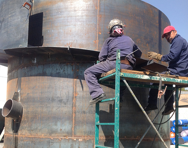 Soldadores de TAZA trabajando en la construcción de un gran tanque de acero. Se observa el proceso de soldadura en la parte superior del tanque.