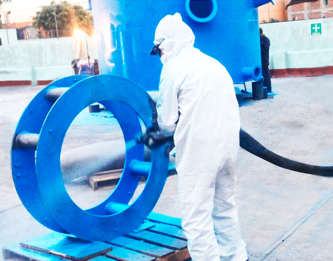 Un técnico de pintura industrial con un traje protector blanco, guantes y gafas, aplicando una capa de polímero azul a una gran brida metálica utilizando una pistola de pulverización. La brida está montada sobre un palé de madera en un área de trabajo al aire libre. En el fondo, se ve un gran tanque cilíndrico, también de color azul, que está siendo recubierto.