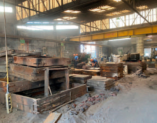 Interior de un taller industrial o fundición, con grandes moldes o cajas de metal, una grúa aérea amarilla en la parte superior y trabajadores con equipo de seguridad en el fondo.