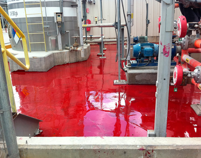 Un piso industrial de color rojo brillante y reflectante, que muestra una capa de polímero recién aplicada. En el área se ven tanques de procesamiento, tuberías, motores y válvulas. El polímero cubre completamente el piso, formando una superficie lisa y resistente.