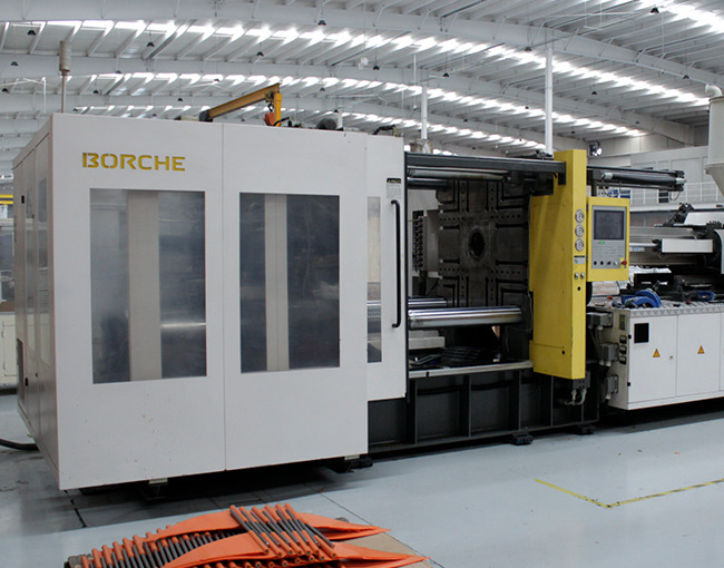Máquina de inyección de plástico de la marca BORCHE en un taller industrial. En el suelo hay piezas terminadas de polímero de color naranja.