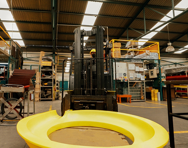 Un montacargas con un operador levanta una pieza de polímero de gran tamaño y color amarillo, con forma de espiral, dentro de un almacén industrial.