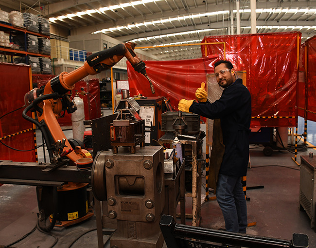 Un trabajador en una fábrica industrial da un pulgar arriba mientras un robot de manufactura KUKA realiza una tarea de producción.