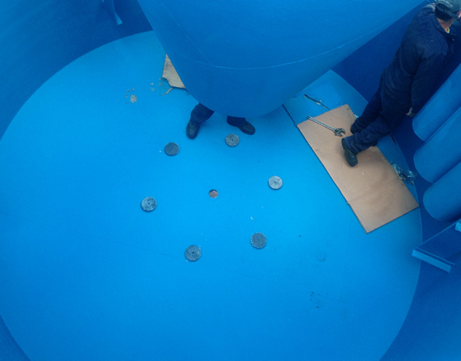 El interior de un tanque industrial circular, recubierto con un polímero de color azul uniforme. Se ven los pies de dos trabajadores sobre la superficie, junto a un conjunto de discos metálicos redondos y un orificio en el centro. La parte inferior de una estructura cónica, también recubierta de polímero, se ve en la parte superior de la imagen.