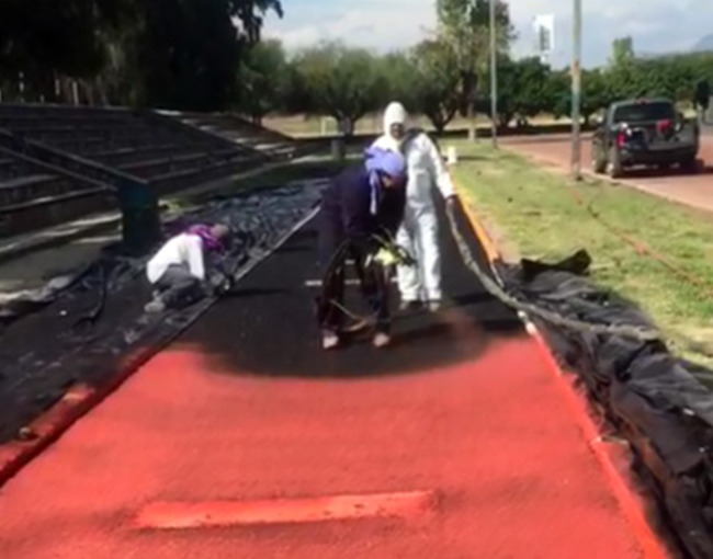 Un equipo de trabajadores, vestidos con trajes protectores, aplicando un recubrimiento de polímero de color rojo a una pista deportiva al aire libre. Un trabajador está arrodillado utilizando una herramienta para esparcir el polímero, mientras otro, de pie, supervisa la aplicación. Las áreas adyacentes a la pista están cubiertas con plástico negro para protección.