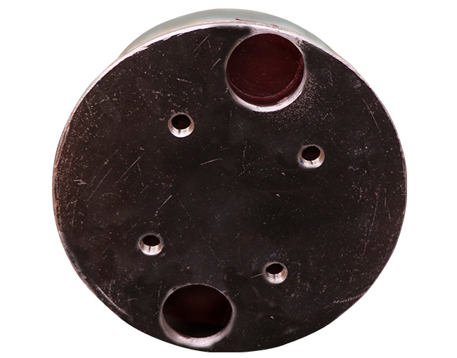 Un tapón o componente cilíndrico de poliuretano de color oscuro, probablemente de poliuretano o un material similar, visto desde arriba. La superficie superior está ligeramente rayada y muestra cuatro orificios más pequeños con rosca, así como dos orificios pasantes más grandes, uno en la parte superior y otro en la inferior, ambos de color rojo oscuro.