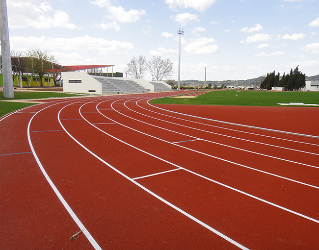 Una moderna pista de atletismo de color rojo ladrillo con carriles blancos. La pista se curva en el primer plano, con un campo de césped verde en el centro. Al fondo, se ven las gradas de un estadio pequeño y edificios entre árboles, con un cielo azul y algunas nubes.