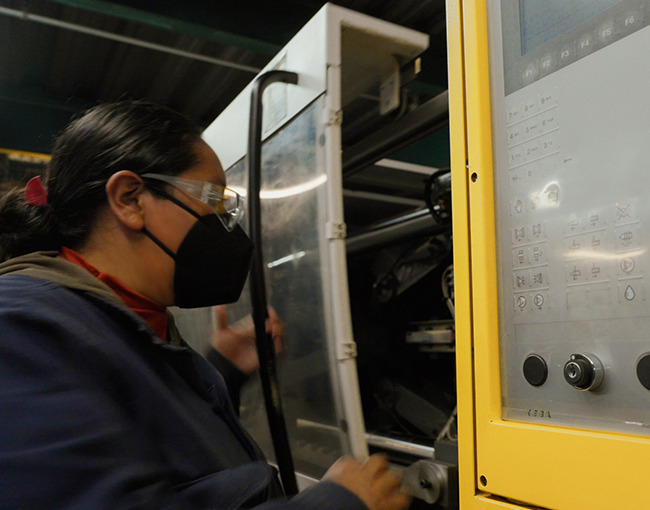 Una trabajadora con equipo de protección personal (mascarilla y lentes) operando una máquina industrial con un panel de control en un taller de fabricación.