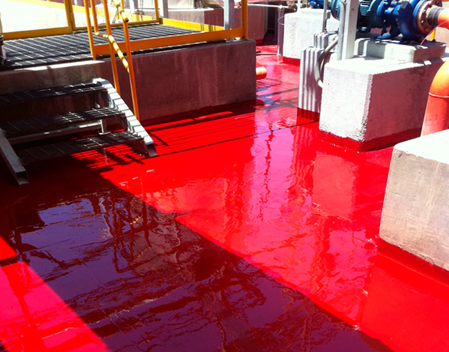 Un piso industrial de concreto recubierto con una capa de polímero de color rojo brillante y reflectante. La imagen muestra la superficie lisa y sellada alrededor de escaleras metálicas, tanques y motores. La luz del sol se refleja en el polímero, destacando su acabado uniforme y liso.