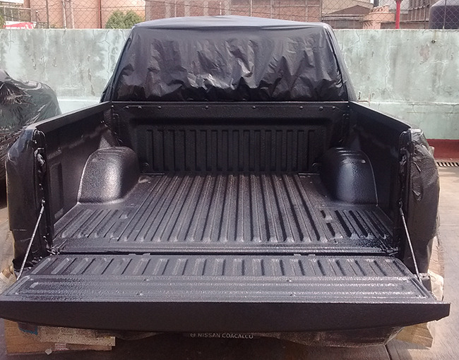 La caja de una camioneta pickup, con la tapa trasera abierta, que muestra un recubrimiento de polímero negro recién aplicado. La superficie del recubrimiento tiene una textura granular, ofreciendo un acabado antideslizante y protector. La cabina y otras partes de la camioneta están cubiertas con plástico negro para protegerlas durante el proceso de aplicación del polímero.
