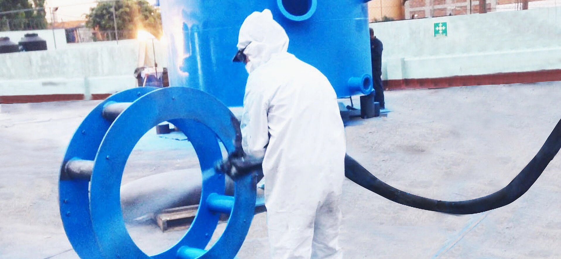 Un técnico de pintura industrial con traje protector blanco, guantes y gafas, aplicando pintura azul a una brida metálica de gran tamaño utilizando una pistola de pulverización. En el fondo, se ve un tanque de tratamiento de agua o contenedor cilíndrico de color azul.