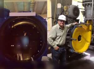 Un técnico con casco de seguridad se encuentra en una planta de producción, posando junto a una gran pieza de maquinaria industrial que ha sido parcialmente desmontada. El componente circular es de color amarillo y está protegido por una carcasa de color azul. El lado izquierdo de la imagen muestra un primer plano del interior de la maquinaria, mientras el lado derecho muestra al técnico con la pieza.