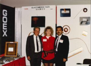 Fotografía de época de tres personas, dos mujeres y un hombre, posando frente a un stand de exhibición con el letrero "ELASTOMEROS TAZA S A DE C V", lo que representa un momento en la historia de la fundación de la empresa.